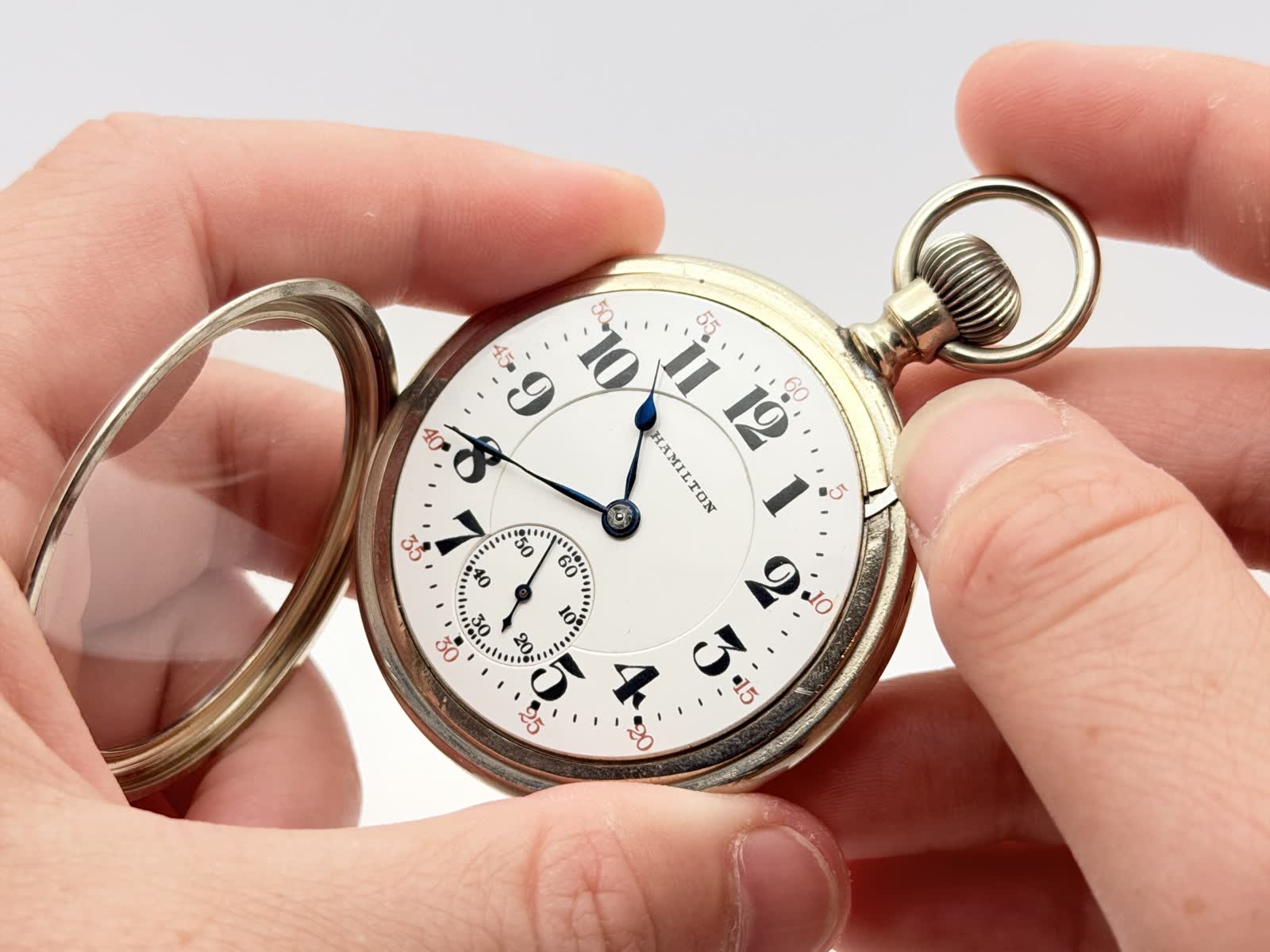 Hands holding a Hamilton pocket watch with the bezel and crystal lifted away, the bare dial exposed