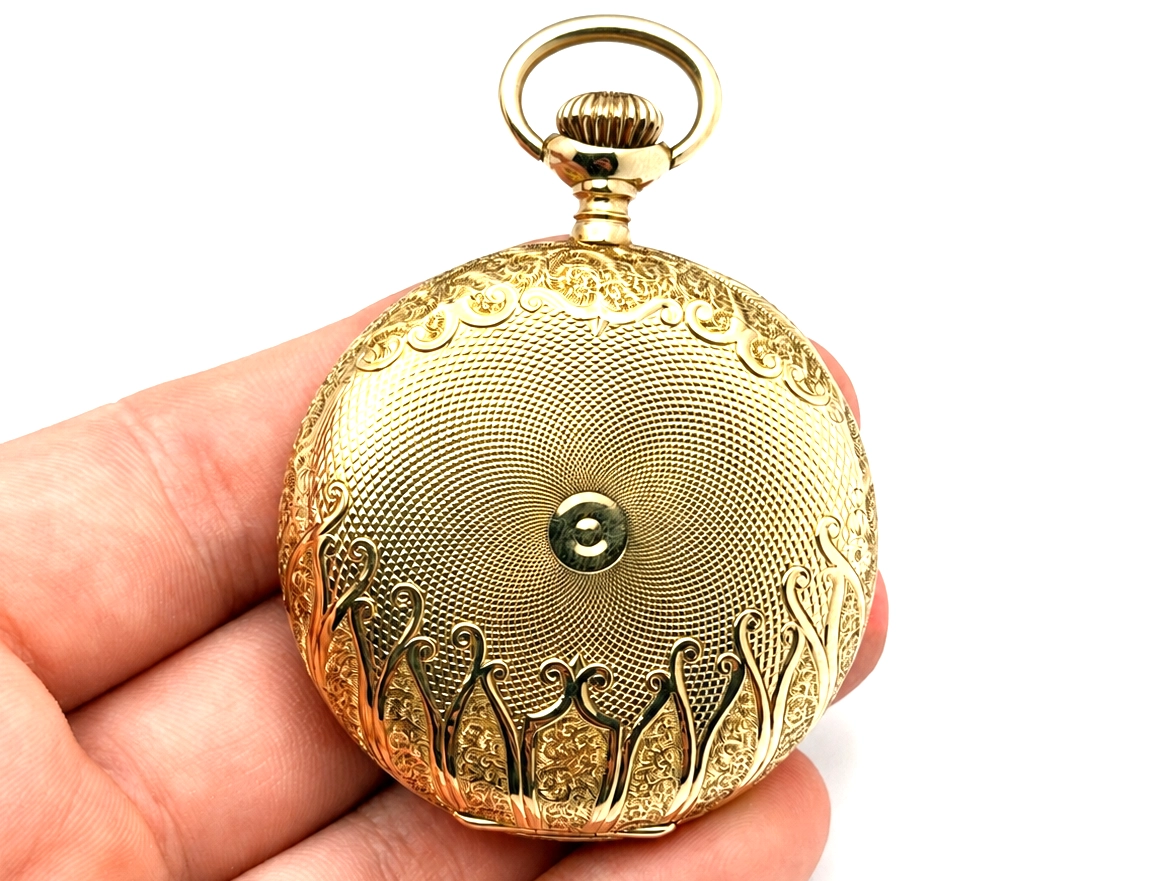 Ornate gold case back with engraved filigree scrollwork and center jewel, displayed in hand.