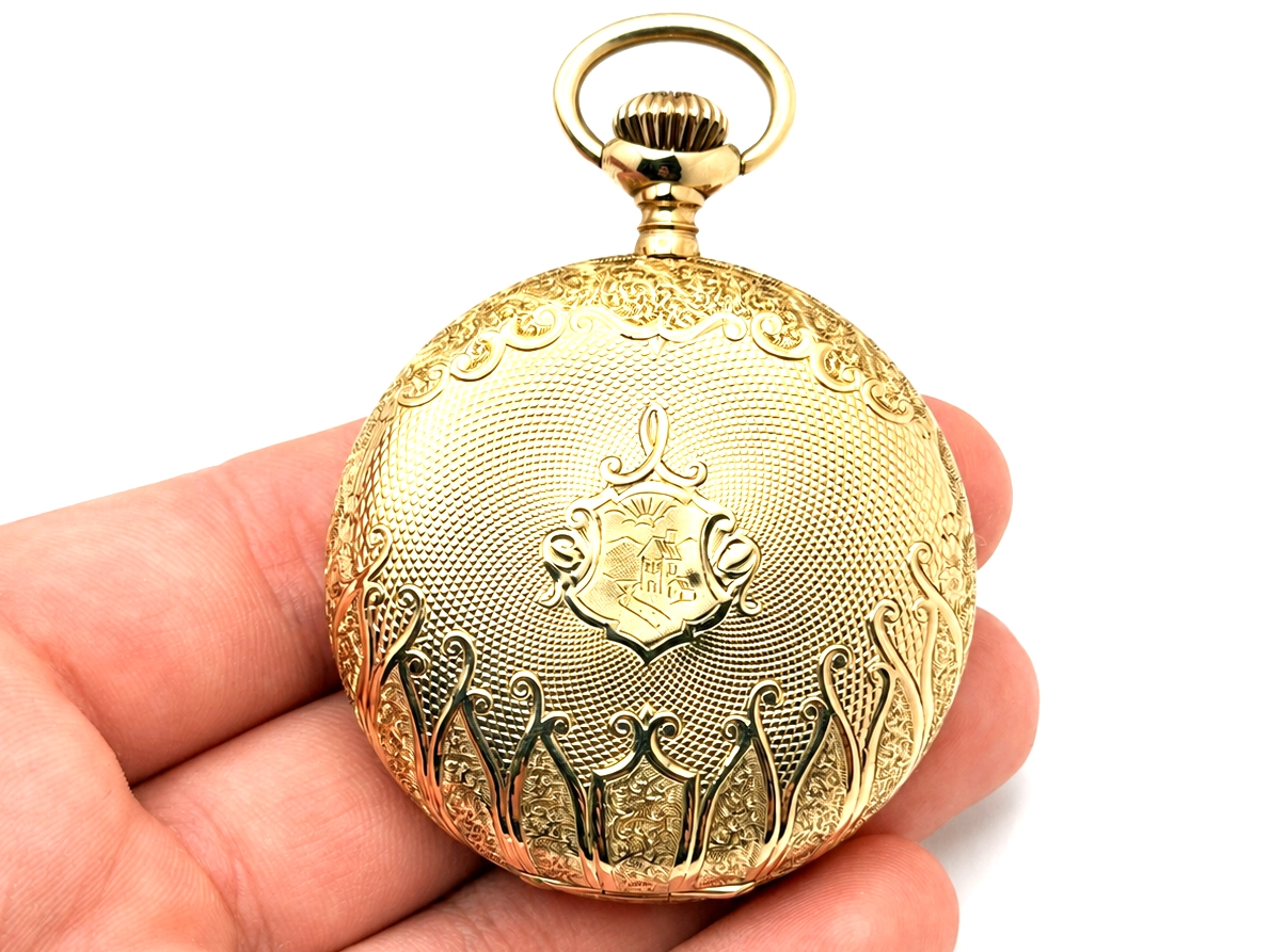 Gold pocket watch case with ornate scrollwork engraving, shield crest, and bow loop held in hand.
