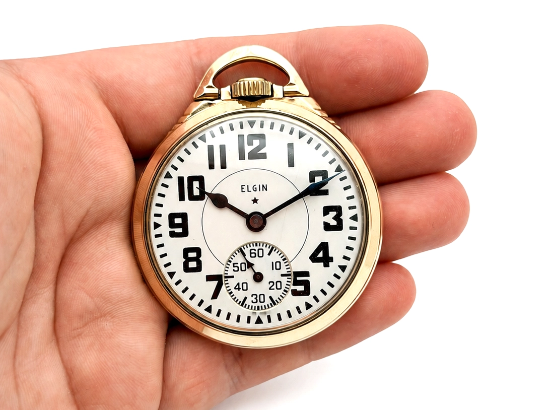 Gold-tone pocket watch with white dial, Arabic numerals, and subsidiary seconds dial held in hand.