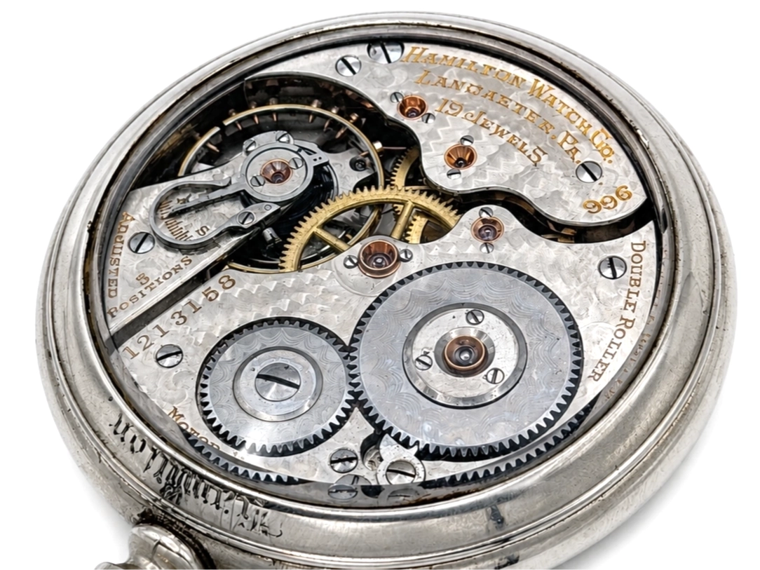 Open movement showing layered gear train with brass gears, jeweled bearings, and Hamilton hallmarks on silvered plate.