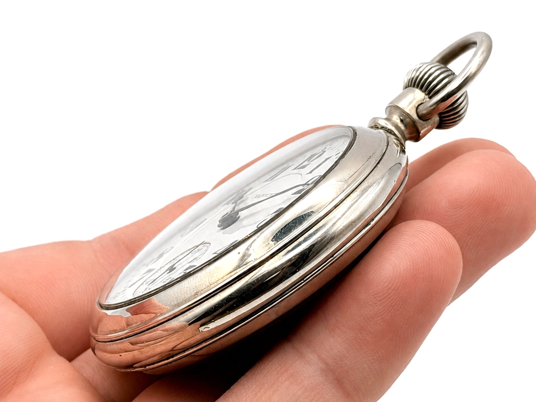 Open pocket watch case with clear crystal held in hand, showing polished metal case with ribbed bow.