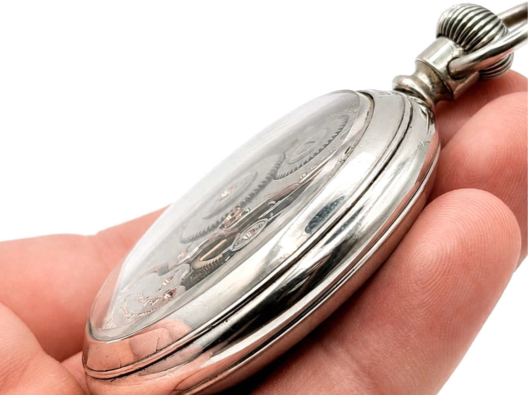 Silver pocket watch case held open showing decorative engraved inner cover and spiral crown.