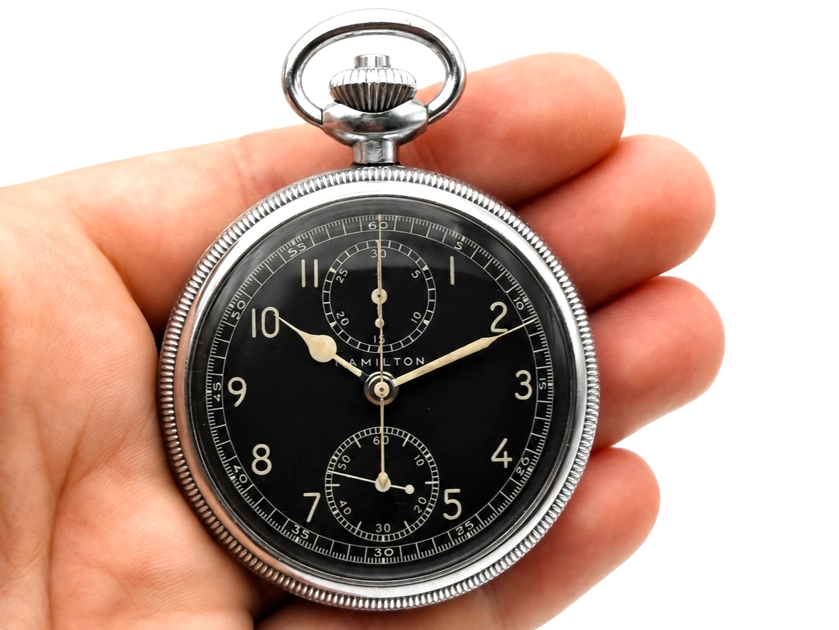 Open pocket watch with black dial, Arabic numerals, chronograph subdials, and luminous hands in hand.