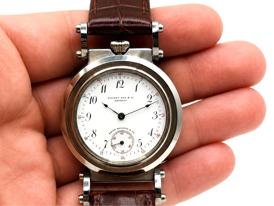 Stainless steel watch with white dial, subsidiary seconds subdial, Arabic numerals, and dark leather strap held in hand.