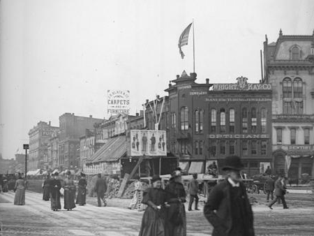 Historic black and white photograph of a crowded city street with period buildings and a flag.