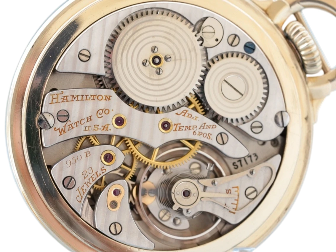 Open pocket watch movement showing gears, jewels, and engraved Hamilton Watch Co. USA plate with temperature and time compensation.