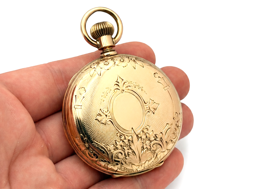 Gold pocket watch case back with ornamental engraving and central cartouche, held in hand.