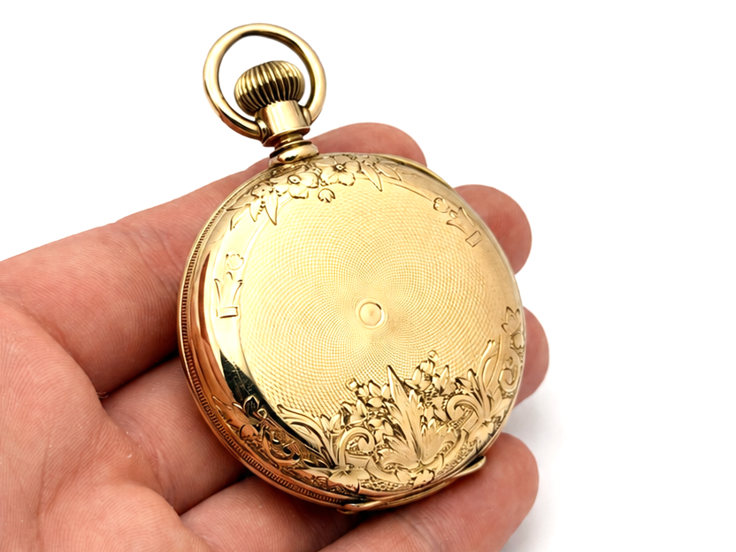 Gold pocket watch case back with ornate engraved floral scrollwork border and plain center panel.