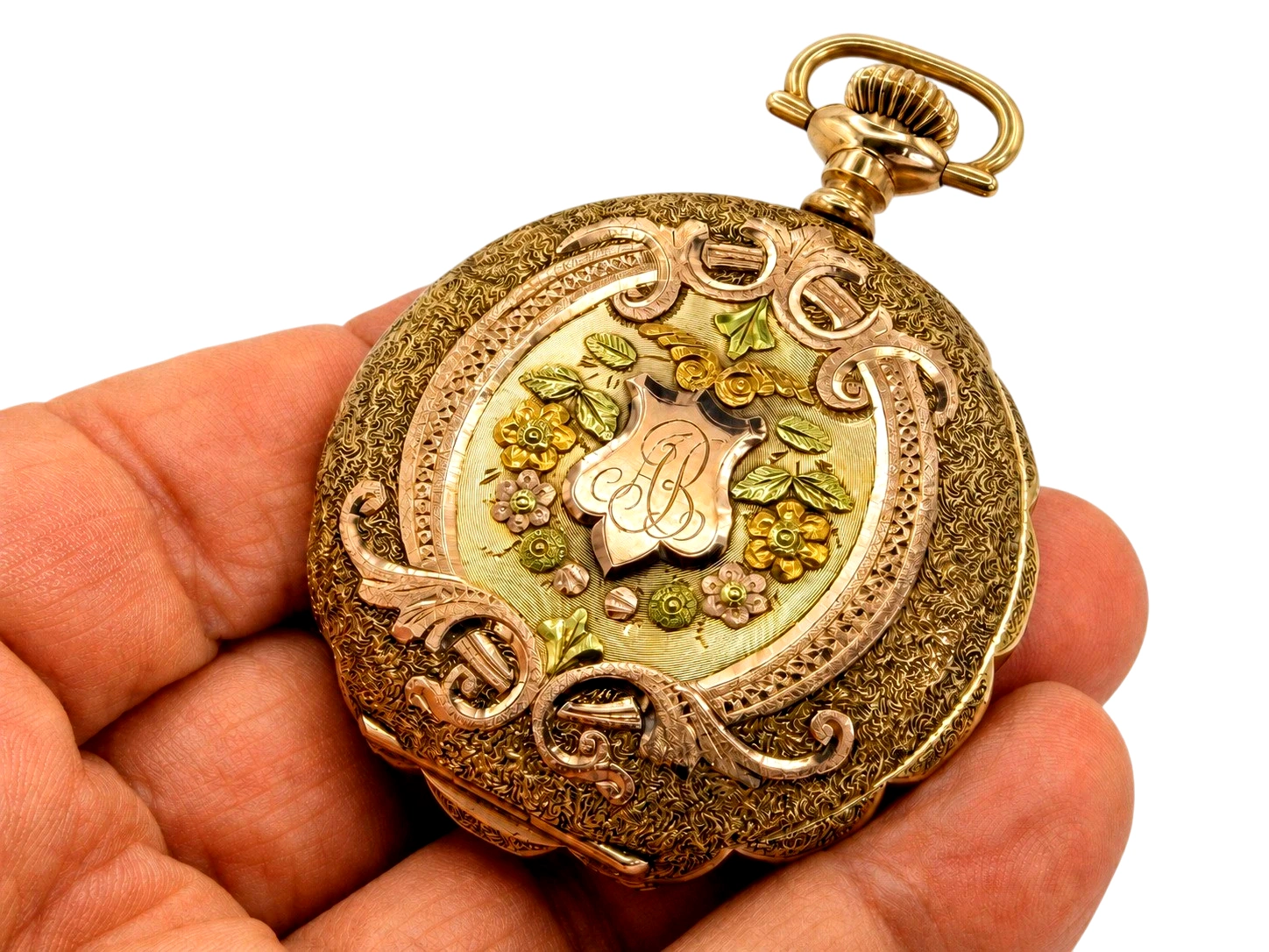 Gold pocket watch case with ornate floral engraving and scrollwork held in hand, case closed.